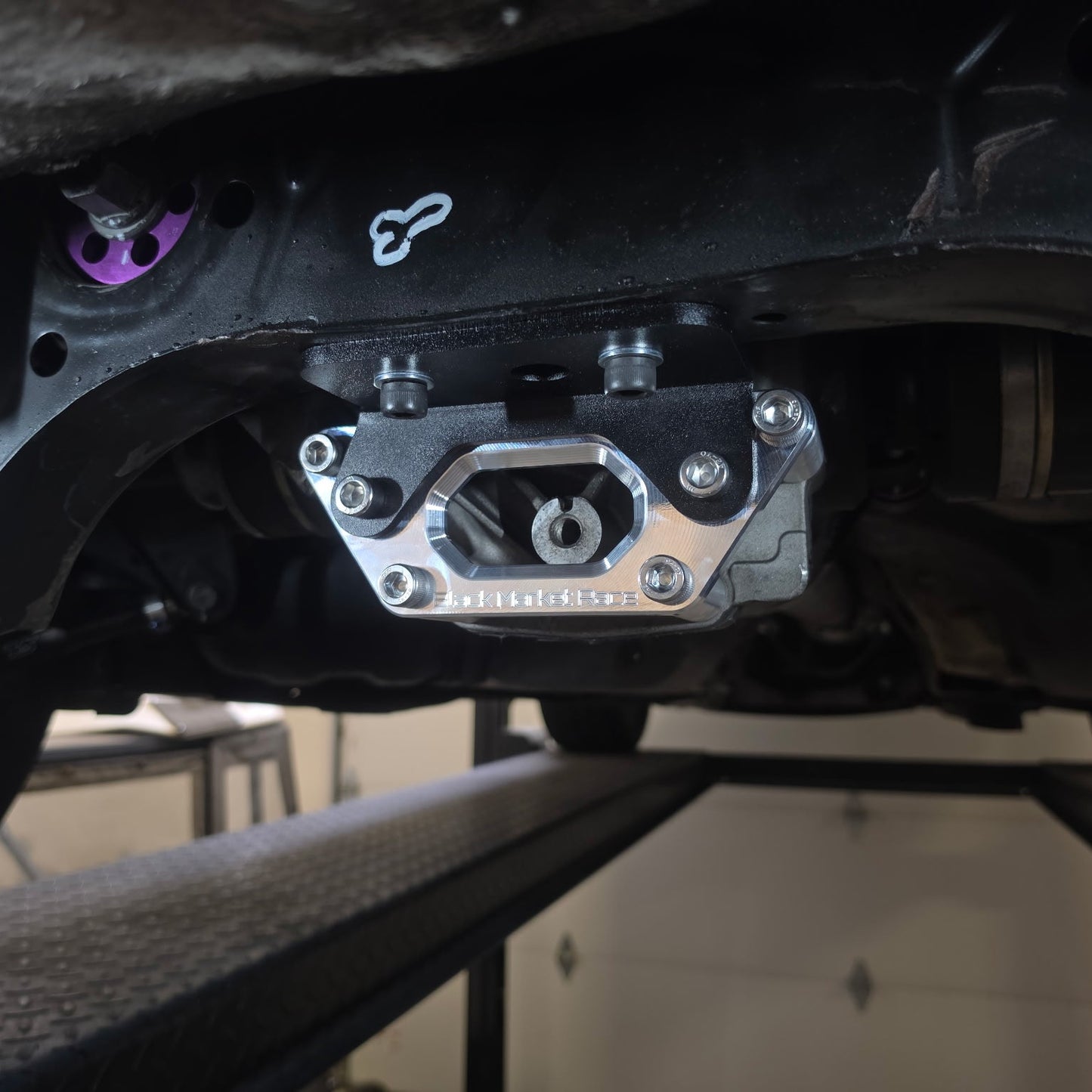 A close-up shot of a metallic silver and black automotive undercarriage component, specifically a chassis bracket for BMW 340i X Drive, mounted on a vehicle lift in a garage setting.